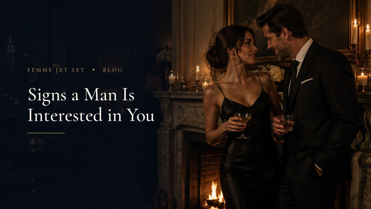 Elegant woman in a black silk slip dress and a man in a dark suit leaning toward her by a candlelit marble fireplace, Signs a Man Is Interested in You cover